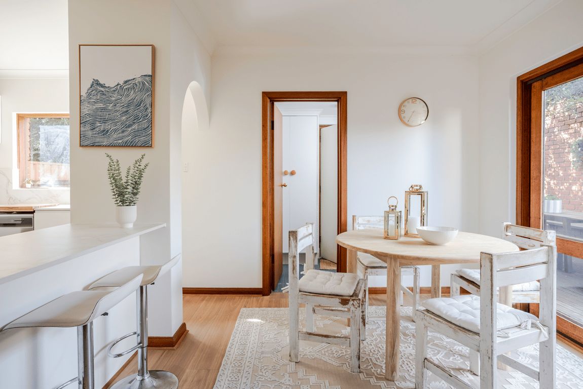 Un comedor con una mesa de madera, sillas blancas y un tapete blanco. Se observa una zona de cocina con armarios blancos y un taburete de bar. En la pared hay una obra de arte enmarcada, y un reloj también es visible.