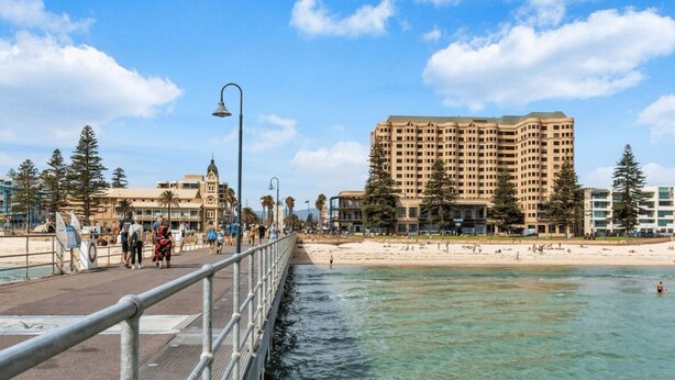 A coastal promenade with a beach, a high-rise building, and a pedestrian walkway. - Belle Escapes - Glenelg Urban Escape (Holdfast Bay)