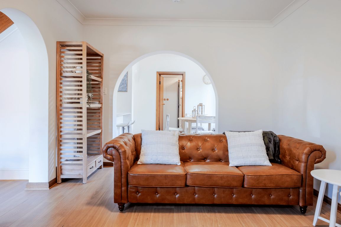 A brown leather sofa with two pillows, a wooden shelving unit, and a white table in a room with wooden flooring and white walls.