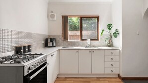 A kitchen with white cabinets, a gas stove, a window with a view of greenery, and a potted plant. - Belle Escapes - Glenelg Urban Escape (Holdfast Bay)