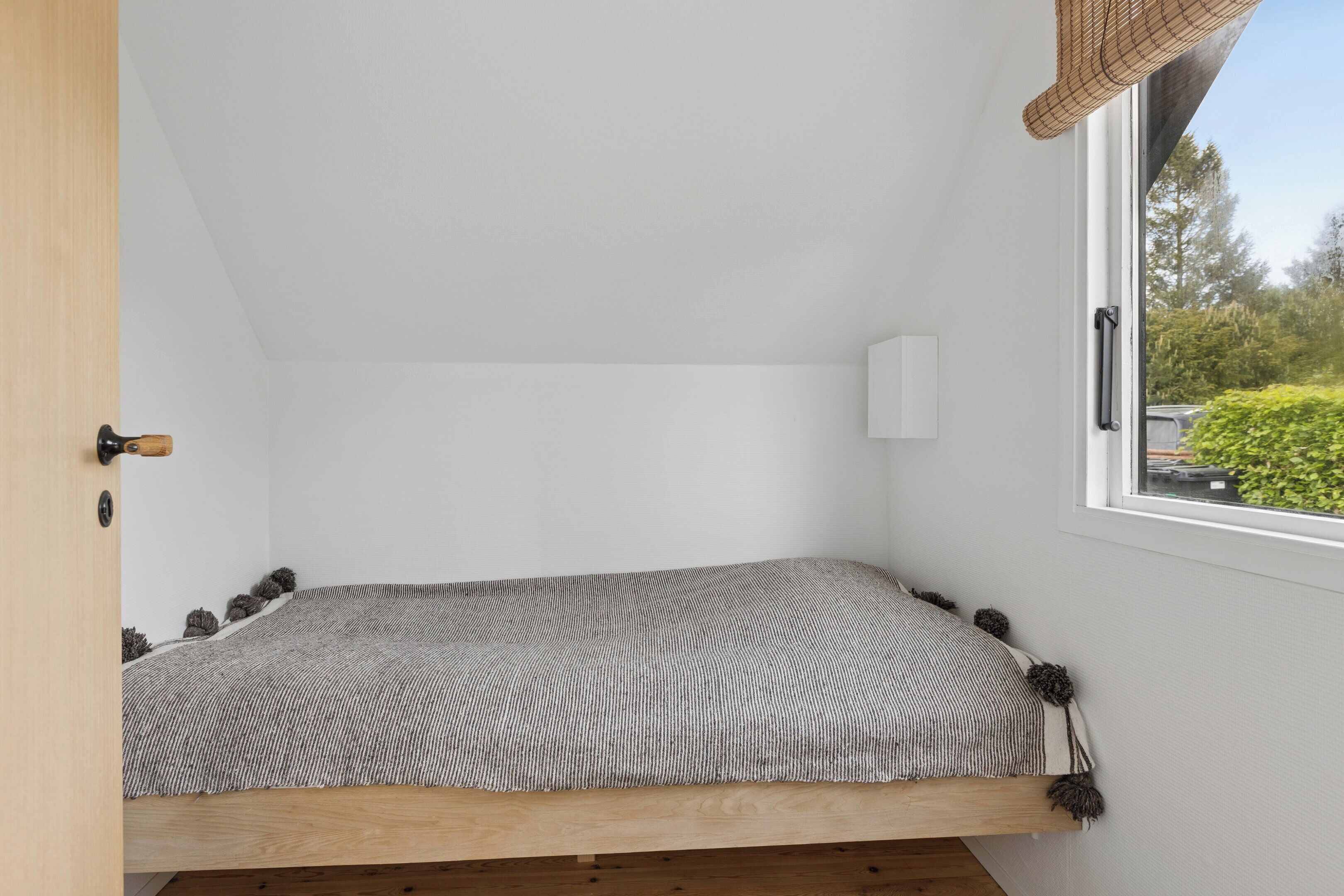 Una habitación pequeña con cama individual, techo inclinado, ventana con vistas a zonas verdes y puerta con manija.