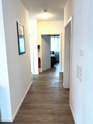 A hallway with wooden flooring, white walls, and a door leading to a room with a bed and a window. - NEW "Nemo" Vacation Rental in Papenburg, Ground Floor, 83 sqm, 2 Bedrooms, Kitchen, Free WiFi, Terrace (Papenburg)