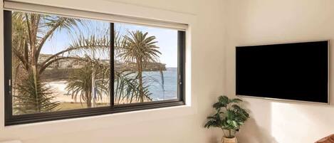 Una habitación con un amplio ventanal con vista a la playa, un televisor de pantalla plana montado en la pared, una mesa de centro redonda y una planta en maceta.