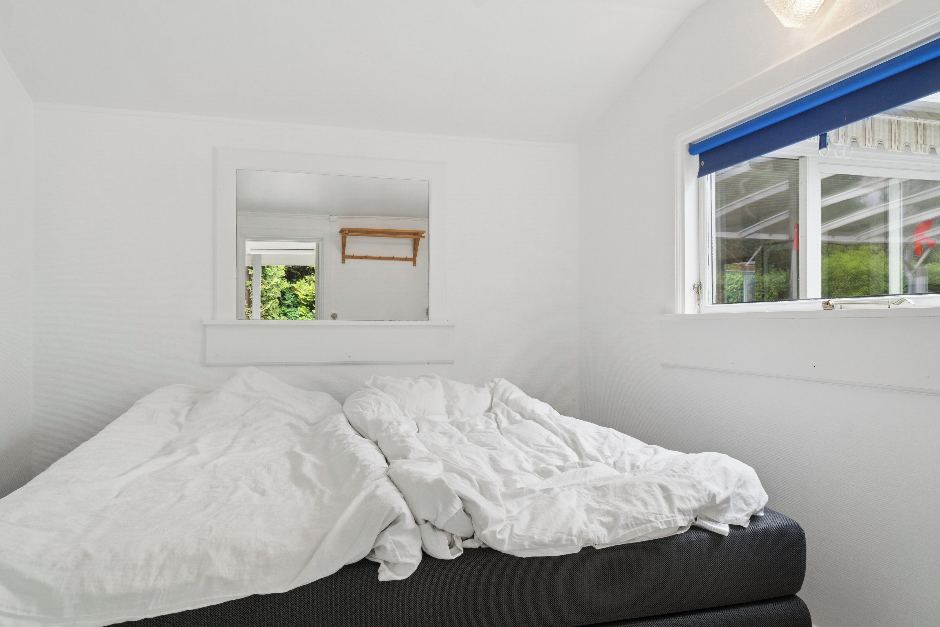 Una cama con ropa blanca, un espejo encima y una ventana con persiana azul.