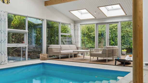 A swimming pool with a wooden deck, outdoor seating, and large windows offering a view of greenery.