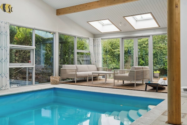 Ein Swimmingpool mit einer Holzterrasse, Sitzgelegenheiten im Freien und großen Fenstern mit Blick auf Grünflächen.
