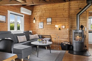Ein gemütliches Blockhauss-Interieur mit einer Couch, einem runden Tisch mit Flaschen und Gläsern, einem Holzofen und gerahmten Bildern an der Wand.