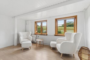 A room with white walls, wooden flooring, two white armchairs, a small round table, and large windows with wooden frames. - Lovely holiday home by the Baltic Sea (Gudhjem)
