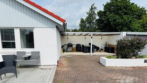 A covered patio with outdoor furniture, a barbecue grill, and a white building with a red roof.