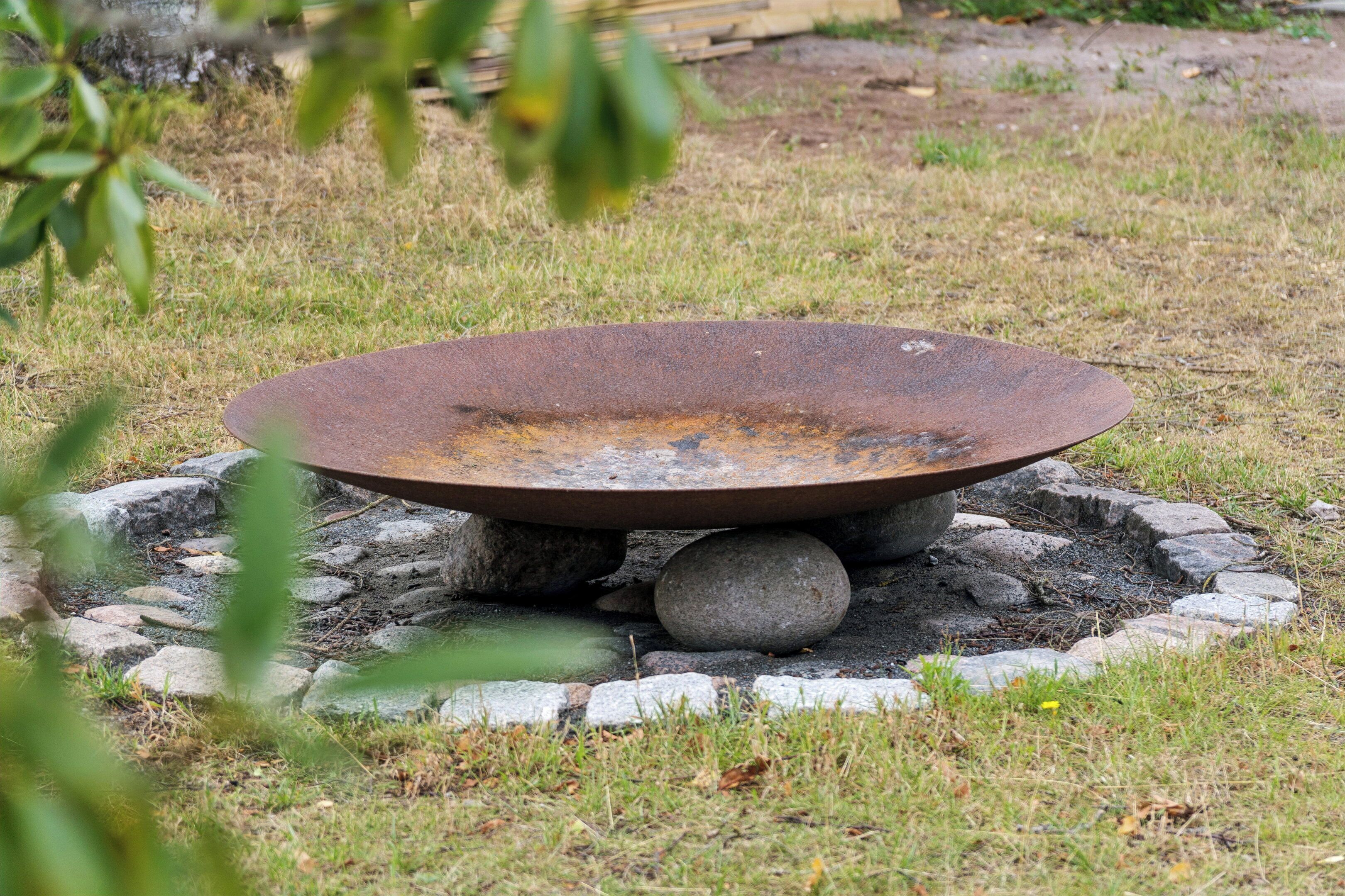 A circular outdoor fire pit with a rusted surface, surrounded by rocks and grass.