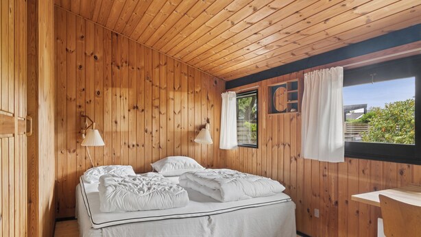 Una habitación con paneles de madera, una cama, dos lámparas de noche, un ventanal con cortinas y una pequeña mesa.