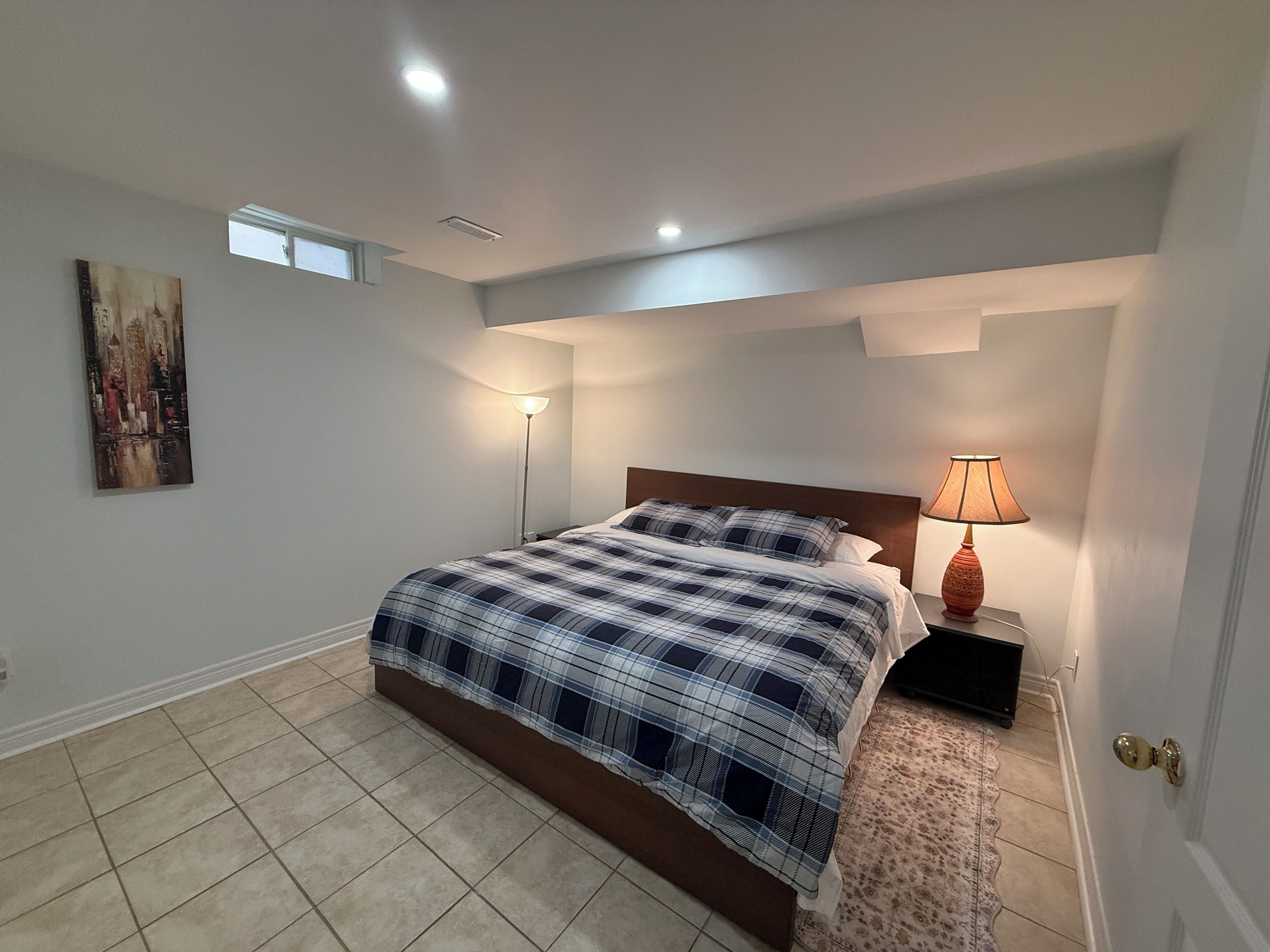 Une chambre à coucher avec un lit, une table de chevet, une lampe, un tableau et une fenêtre.
