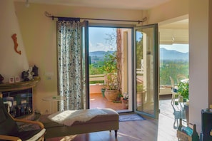 A room with a sofa, a small table, a fireplace, and a sliding glass door leading to a balcony with plants and a view of the countryside. - Villa Rodi - 3 Bedroom Holiday Home (Zakynthos)