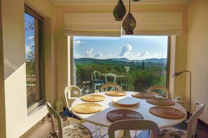 A dining area with a table set for four, chairs, a view of greenery and hills, and a window with a view. - Villa Rodi - 3 Bedroom Holiday Home (Zakynthos)