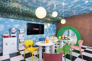 Una habitación con piso a cuadros, televisor montado en la pared, máquinas expendedoras, sillas coloridas y un arco verde.