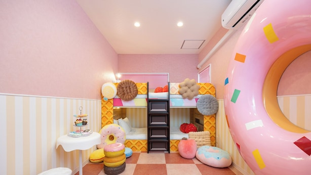 Une chambre d’enfant avec des lits superposés, une grande décoration murale en forme de beignet, une petite table avec un gâteau et une poubelle.