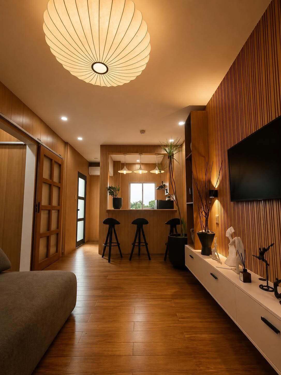 A modern interior with a wooden floor, a sofa, a dining area with stools, a kitchen with a TV, and a ceiling light fixture.