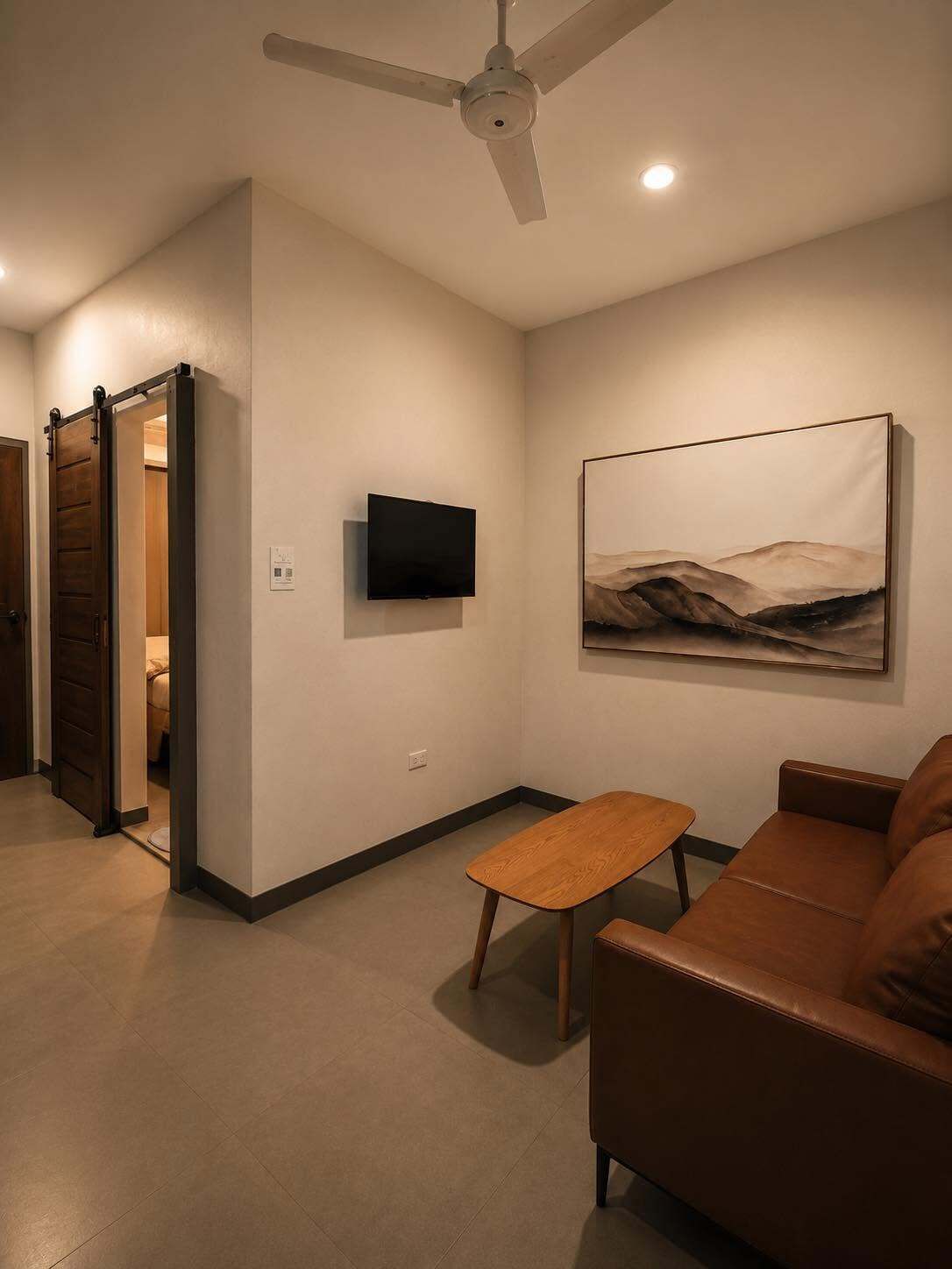Una sala de estar con un sofá de cuero marrón, una mesa de centro de madera, un televisor de pantalla plana montado y un ventilador de techo.