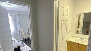 Un pasillo que conduce a un dormitorio y baño, con una cama, una silla y un lavamanos.