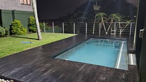 A rectangular swimming pool with a dark wooden deck, surrounded by a low fence and greenery. - La Lucia Home With a View (uMhlanga)