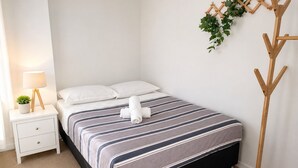 A neatly made bed with striped bedding, a bedside lamp, a small potted plant, and a wooden coat rack with greenery. - The Sunset Perch | Panoramic River Escape (South Brisbane)