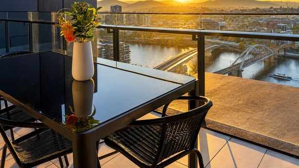 A balcony with a glass table and chairs, a vase with flowers, and a cityscape view at sunset. - The Sunset Perch | Panoramic River Escape (South Brisbane)