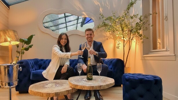 Een modern interieur met een glazen dak, blauwe gestoffeerde zitplaatsen en een ronde houten tafel met een fles en glas.