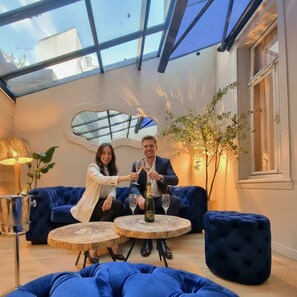 Um interior moderno com teto de vidro, assentos estofados em azul e uma mesa redonda de madeira com uma garrafa e um copo.