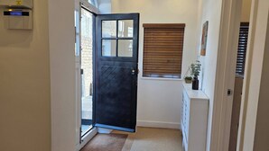 A hallway with a black door, wooden flooring, a white cabinet, a window with blinds, and a wall-mounted light fixture. - Boutique Dairy Conversion Near Ilford Station (Ilford)