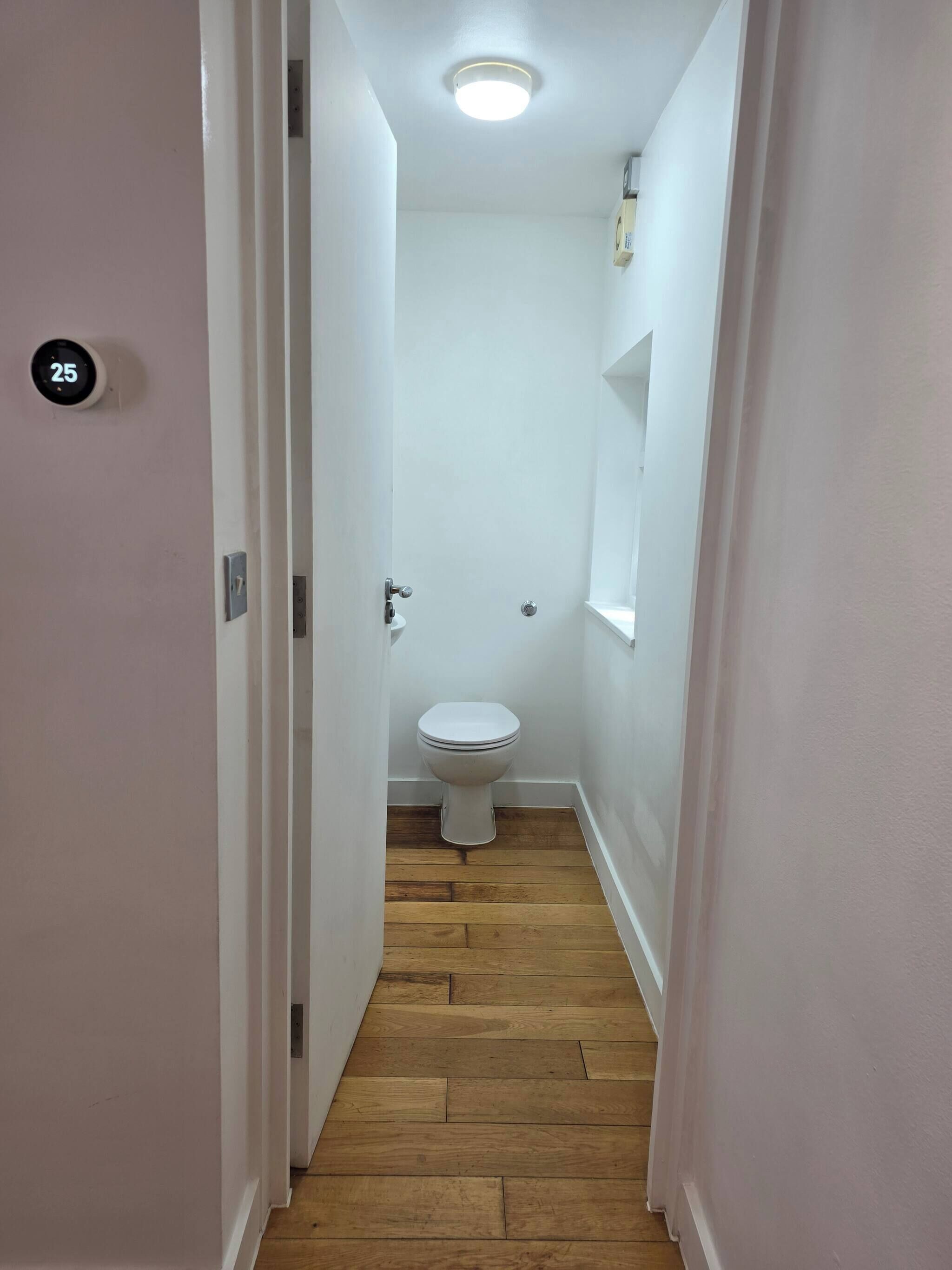 A narrow hallway leading to a bathroom with a white toilet, wooden flooring, and a ceiling light.
