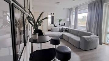 A modern living room with a grey sofa, black bar stools, a potted plant, and a dining area with a table and chairs.