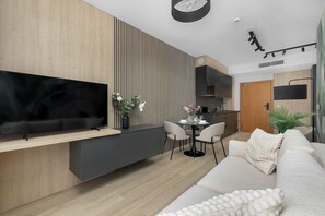 A modern living room with a flat-screen TV, a sofa, a dining area with chairs, and a kitchen in the background. - Comfortable Apartment with Air Conditioning and Parking | Close to the Beach (Mielno)