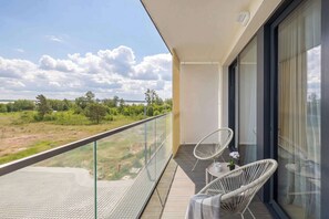 Ein Balkon mit Glastrennwand, zwei Korbstühle, ein kleiner Tisch mit Blume und Blick auf eine Rasenfläche und Bäume.