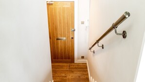 Una puerta de madera con pomo y placa metálica, una escalera con escalones de madera y barandillas blancas, y un pasamanos metálico en la pared.