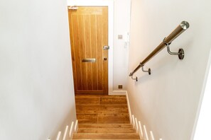 Eine Holztür mit Griff und einer Metallbeschriftung, eine Treppe mit Holzstufen und weißen Geländern sowie ein Metallhandlauf an der Wand.