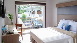 A bedroom with a bed, a desk with a chair, a plant, and a balcony with chairs. - Adela Resort Panglao (Panglao)