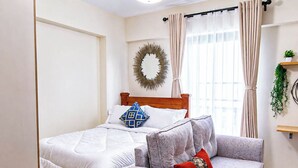 Una camera da letto con un letto, una poltrona, un tavolino, una mensola a parete, una tenda e una lampada a sospensione.