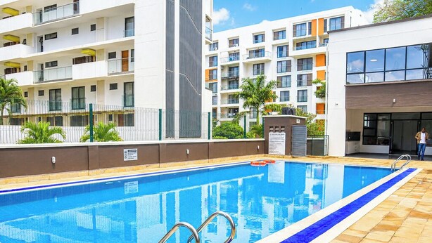 Una piscina con ringhiere in metallo, circondata da un solarium piastrellato, adiacente a un edificio a più piani con balconi e finestre.
