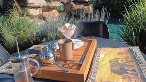 A table with a coffee pot, a mug, a tray, and a plate with food, set outdoors with a pool and rocks in the background.