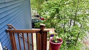 A wooden deck with a black mat, a brown railing, and potted plants with flowers. - The Birch Loft - Woodland Retreat (Anchorage)