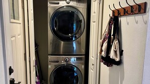 A laundry room with two stacked front-loading washing machines, a sign above the door, and a coat rack with hanging items. - The Birch Loft - Woodland Retreat (Anchorage)
