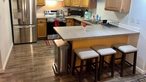 A modern kitchen with stainless steel appliances, wooden cabinets, a granite countertop, and bar stools. - The Birch Loft - Woodland Retreat (Anchorage)