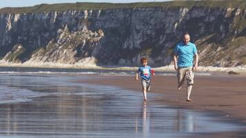 Uma praia com uma pessoa e uma criança caminhando na areia, falésias ao fundo.