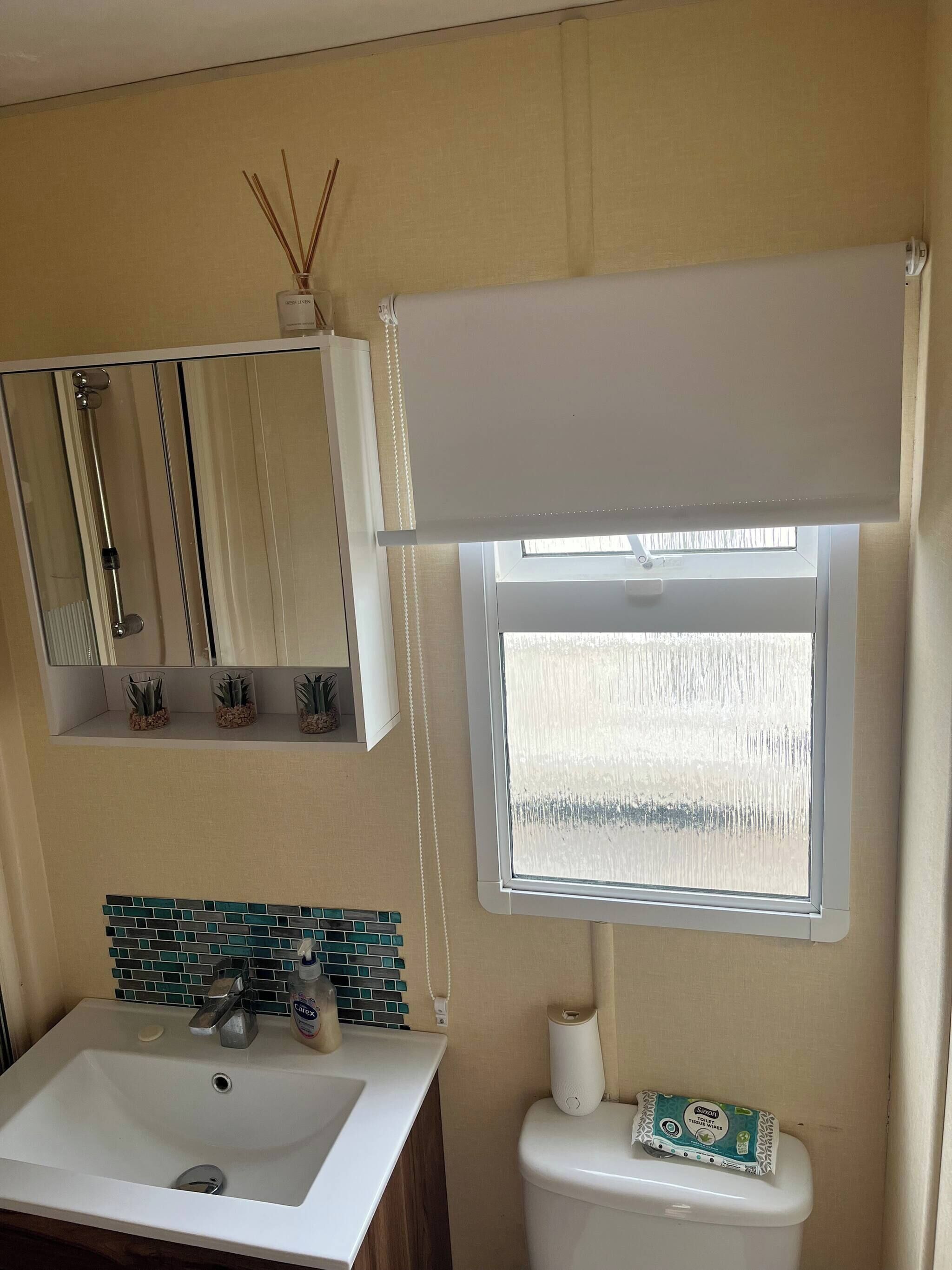 A bathroom with a sink, mirror, toilet, and window with a blind.