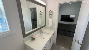 A modern bathroom with a large mirror, two sinks, a vanity, a TV mounted above the sink, and a door leading to another room. - Chris and Carmen Place (Mission)