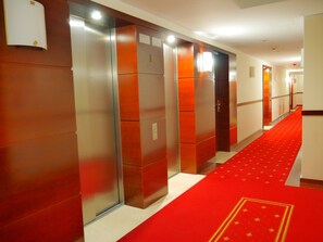 Un pasillo de hotel con ascensores, alfombra roja y pared blanca.
