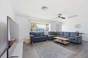 A modern living room with a sectional sofa, a flat-screen TV, a ceiling fan, and a large window.