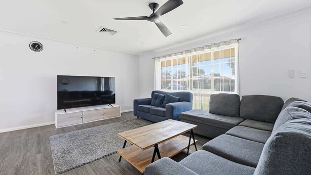 Ein modernes Wohnzimmer mit Flachbildfernseher, Deckenventilator, großem Fenster und grauem Ecksofa.