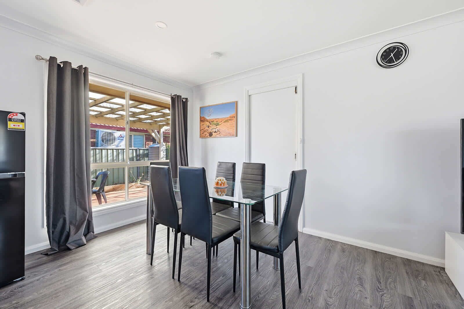 A dining area with a glass table and four chairs, a door with a clock, and a window with curtains.