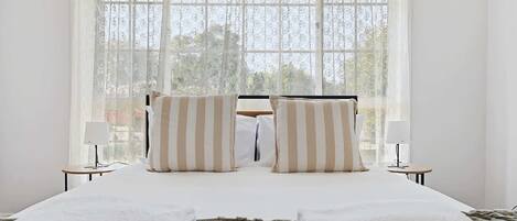 Uma cama com duas almofadas, uma mesa de cabeceira com abajur e uma janela com vista para árvores.